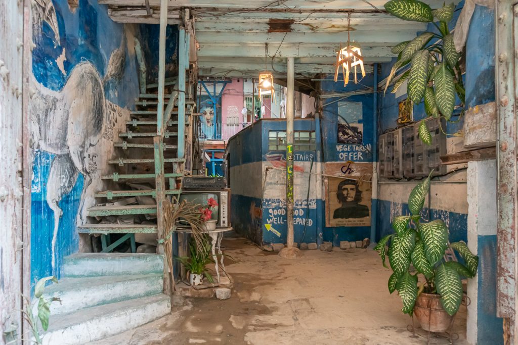 Photo from Havana, Cuba showing a bit of a rundown interior with a staircase, art and posters on the wall, and a couple of plants.  
