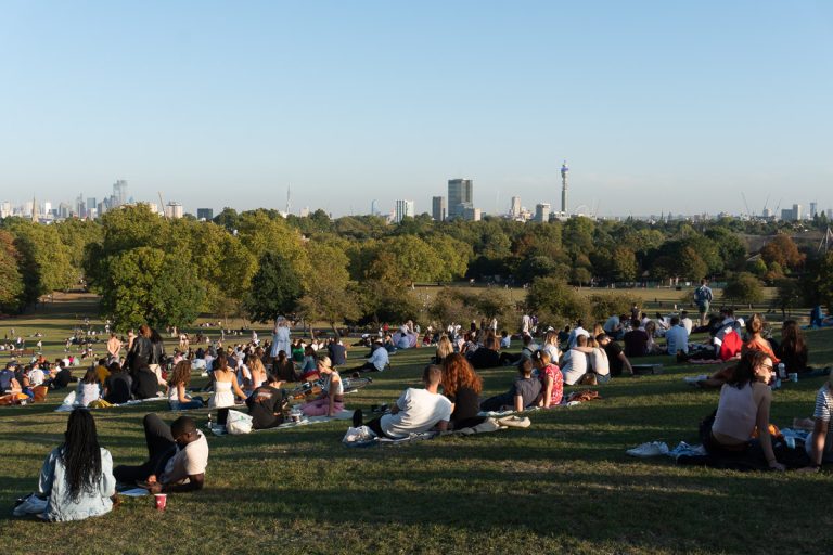 A wide shot of a crowded green grassy hill at Primrose Hill in London during golden hour, with groups of people picnicking and the city skyline visible in the distance.