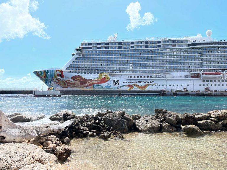 The Norwegian Getaway cruise ship docked at the Aruba port, showing its colorful hull art above clear turquoise water, viewed from a rocky shore on a sunny day.