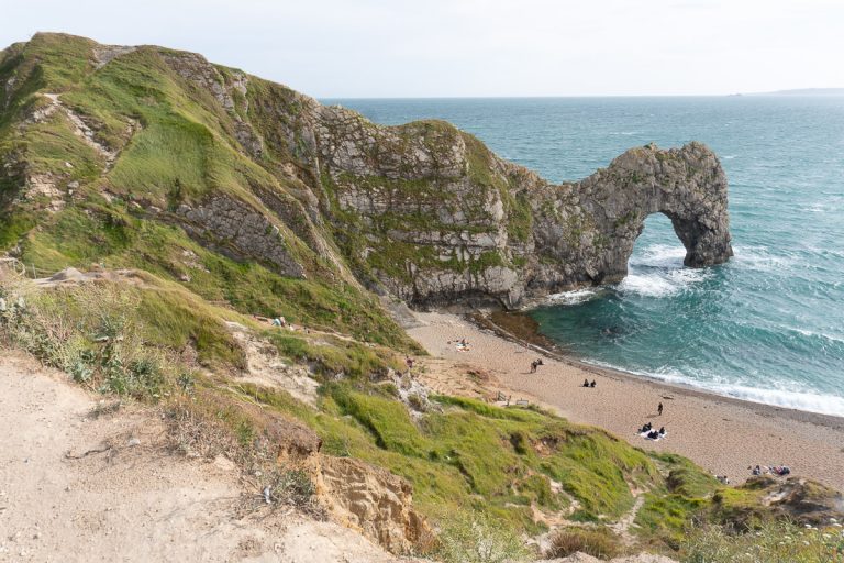 Looking down from a grassy cliffside path at the iconic Durdle Door natural stone arch jutting into the turquoise ocean with a sandy beach below in Dorset, England.
