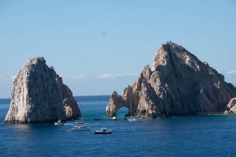 The iconic El Arco natural limestone arch rock formation at Land's End in Cabo San Lucas, Mexico, surrounded by deep blue ocean water with boats and yachts sailing nearby under a clear sky.