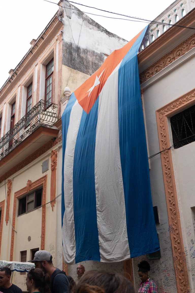 A massive Cuban national flag drapes down the side of a historic, weathered colonial building over a crowded street in Old Havana.