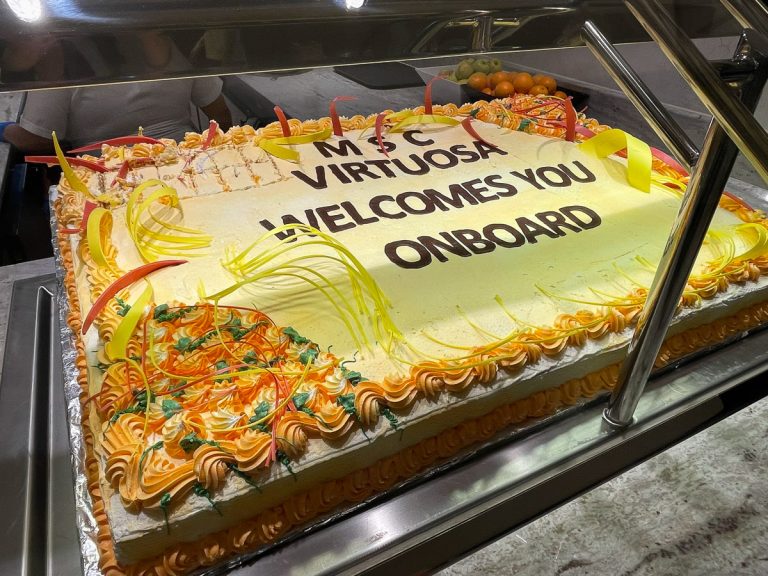 A large decorated sheet cake with orange and yellow frosting piping that reads "MSC Virtuosa Welcomes You Onboard" on display at the cruise ship buffet.