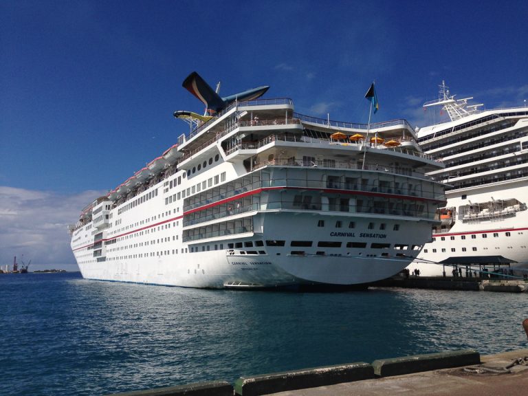 The aft view of the Carnival Sensation cruise ship docked at the pier in Nassau, Bahamas, showing its iconic whale-tail funnel and the Bahamian flag under a deep blue sky.
