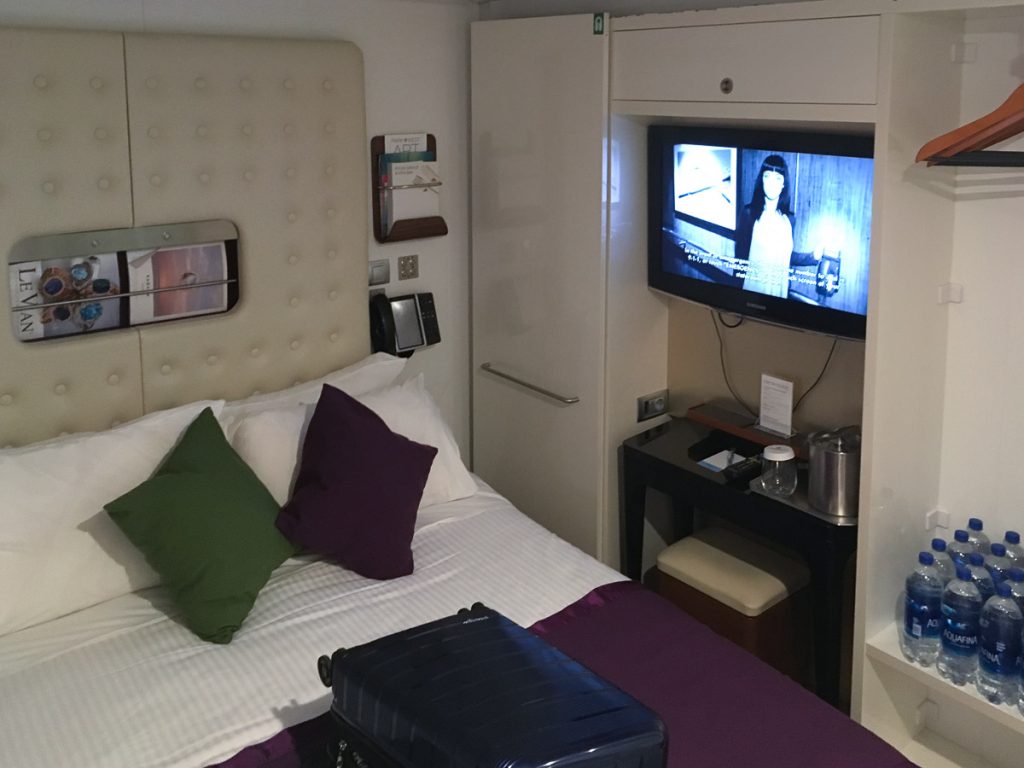 An interior view of a Norwegian Epic cruise ship Studio Cabin for solo travelers, showing a full bed with accent pillows, a flat-screen TV, a small desk, and storage shelving.