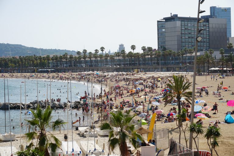 A crowded sandy beach with sunbathers under colorful umbrellas and people swimming in the ocean, with palm trees and modern city buildings in the background on Somorrostro Beach in Barcelona, Spain.