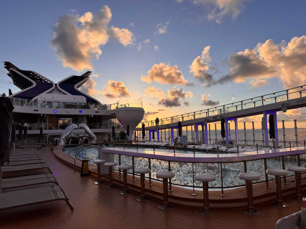 Sunset over the resort deck on the Celebrity Apex cruise ship, featuring an outdoor pool bar, martini glass hot tub, and lounge chairs.