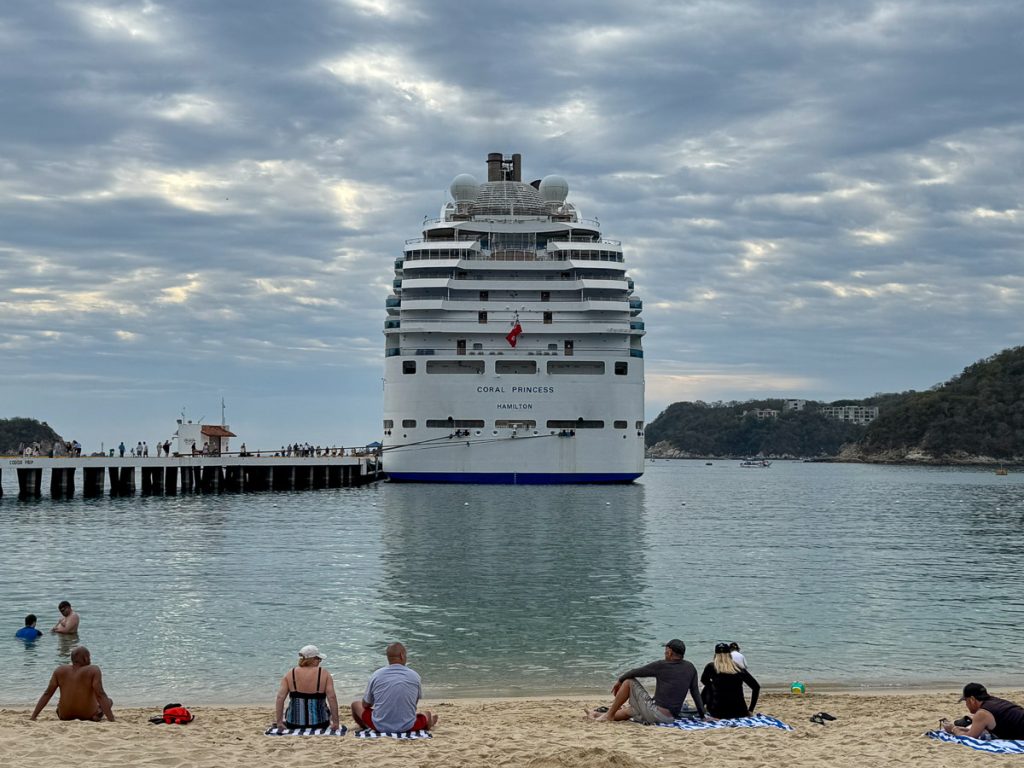 Aft view of the Coral Princess cruise ship at the Santa Cruz Huatulco Cruise Pier with people sitting on the beach in the foreground.