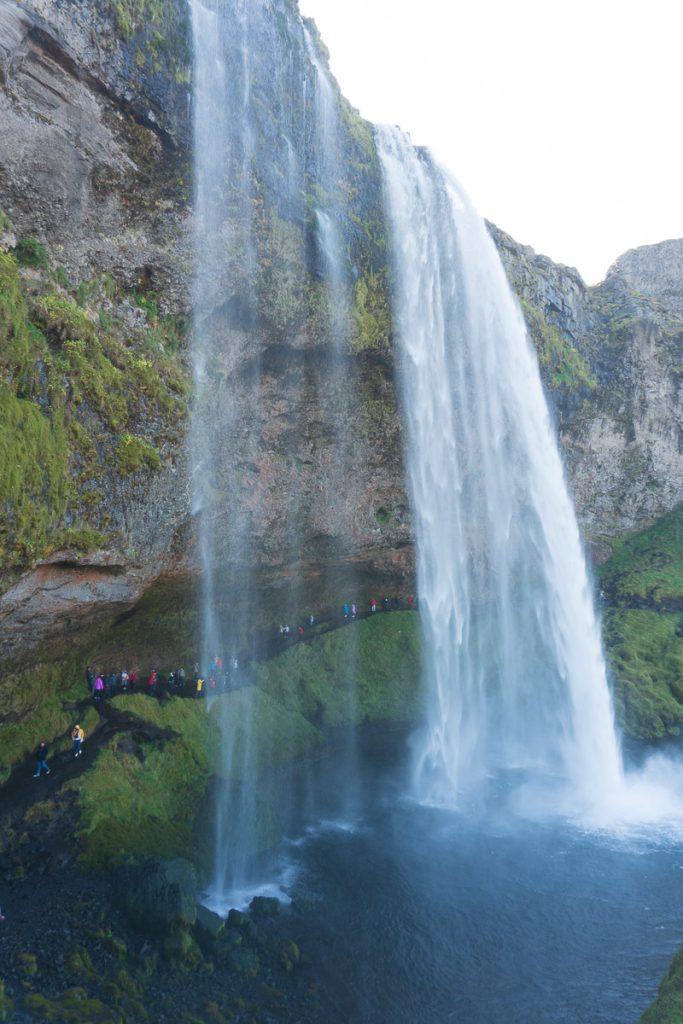 A dramatic wide shot of Seljalandsfoss waterfall in Iceland, showing the rushing water cascading down the cliffside with tourists walking along the rocky path directly behind the falls.