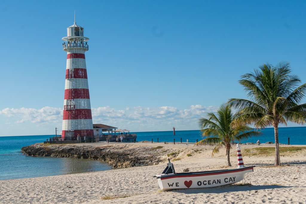 A red and white striped lighthouse on a rocky point at Ocean Cay MSC Marine Reserve, with a white decorative boat reading 'We Heart Ocean Cay' sitting on the sandy beach foreground under a clear blue sky.