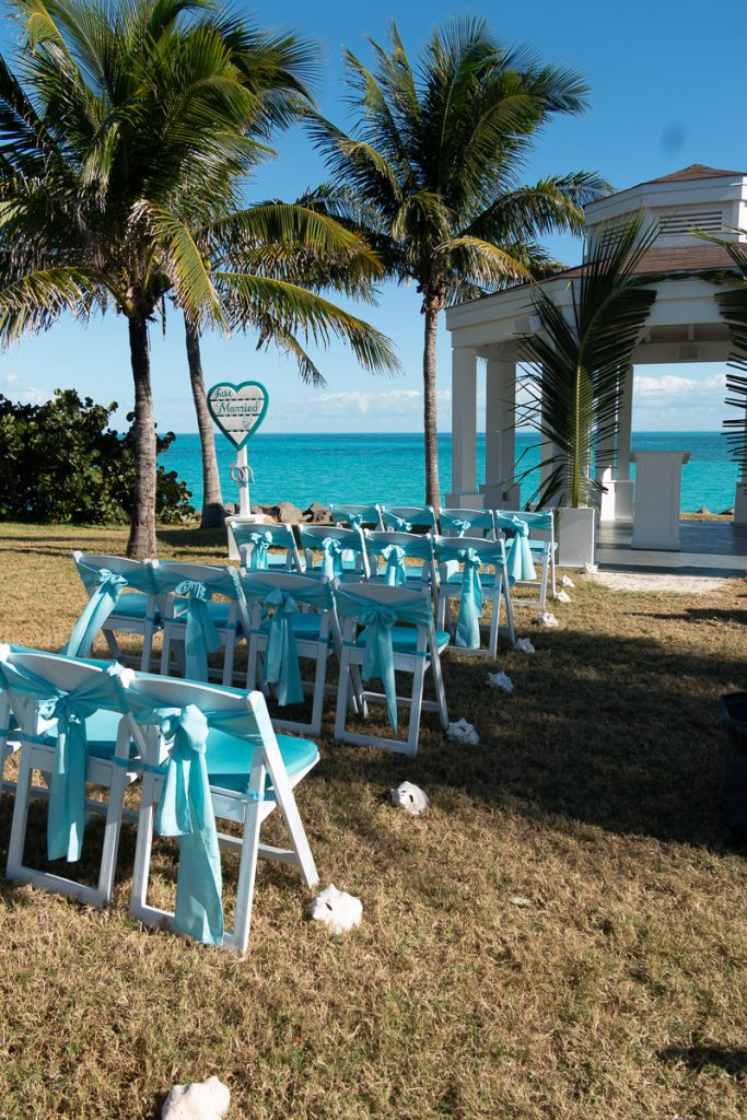 An outdoor wedding ceremony setup on the grass at Ocean Cay MSC Marine Reserve, featuring white chairs with bright turquoise sashes, palm trees, a white gazebo pavilion, and a 'Just Married' heart sign overlooking a clear blue ocean.