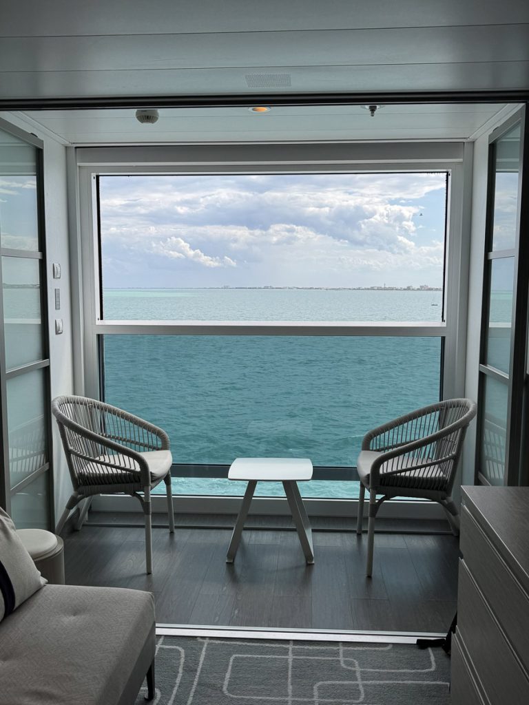 An interior view looking out of a Celebrity Apex Infinite Veranda cabin. Two grey wicker-style chairs with cushions and a small white side table sit in front of the floor-to-ceiling glass panel, which shows clear ocean water and a distant landline under a cloudy sky.