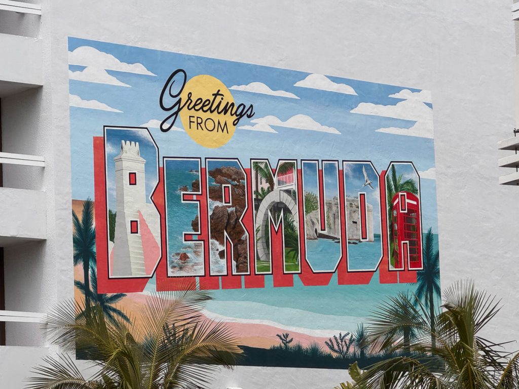 A colorful, large-scale outdoor wall mural on a building in Hamilton, Bermuda, that reads "Greetings FROM BERMUDA." Each of the large block letters in "BERMUDA" contains smaller painted scenes of local landmarks like lighthouses, beaches, forts, and red phone boxes. Palm fronds are visible in the foreground.