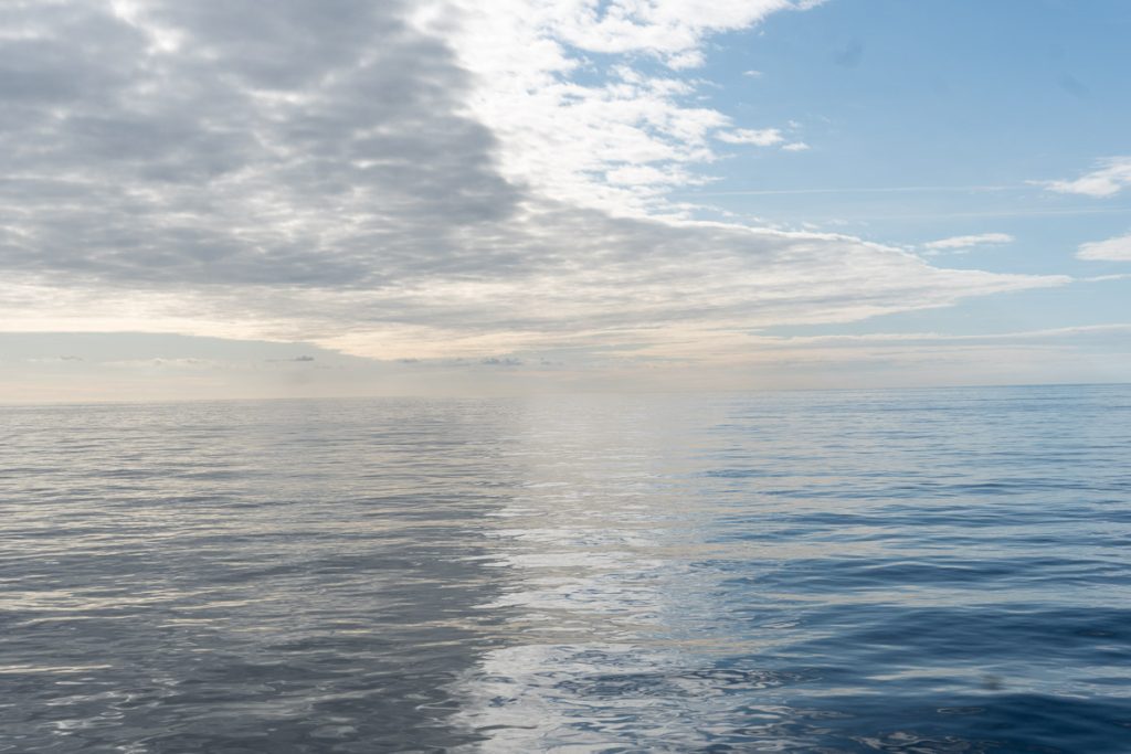 A horizontal photo taken from a ship at sea, showing calm, gently rippling deep blue ocean water under a sky filled with layered white clouds and patches of blue. The water extends to the horizon.