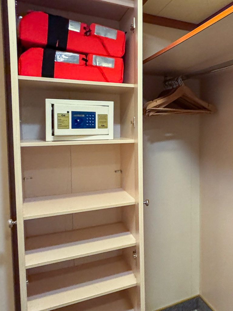 Open cruise ship closet featuring an in-room personal safe on a shelf and bright orange life jackets overhead